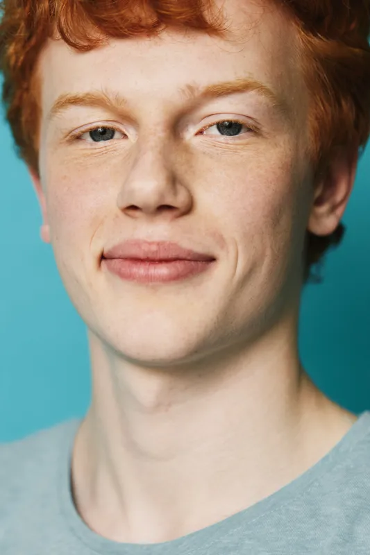 Portrait of young redhead man with freckles smiling with calm expression against blue background wearing casual shirt, face closeup, natural light and relaxed mood.
