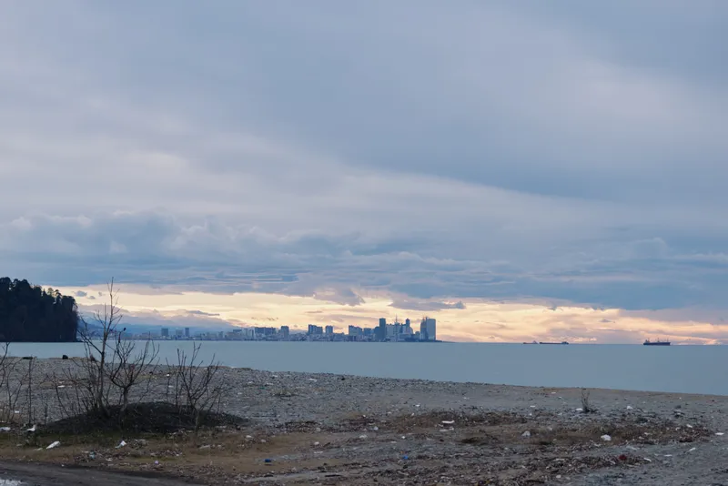 Shoreline with distant city skyline across calm water under an overcast sky, subtle sunset glow on the horizon, rocky beach foreground.