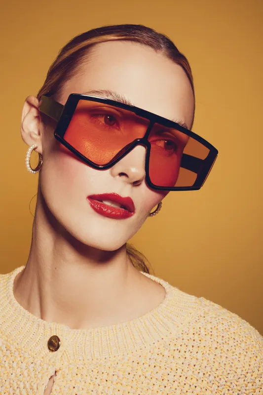 Stylish woman with oversized orange sunglasses and red lipstick poses against yellow background. Fashion portrait with modern eyewear and trendy makeup concept.