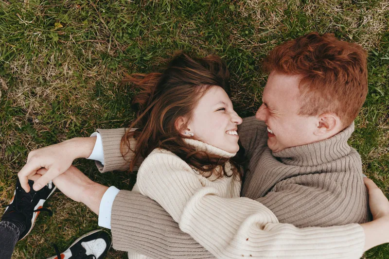 A young couple embraces and smiles warmly while lying on the grass wearing cozy sweaters in a natural outdoor setting, capturing a candid moment of connection and happiness.