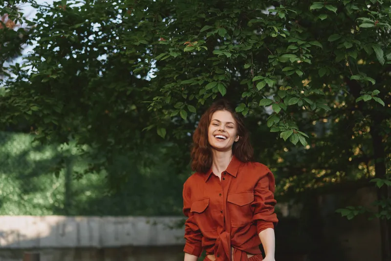 Young woman with curly brown hair smiles brightly in a red shirt outdoors among lush green trees, enjoying a carefree lifestyle moment in a sunlit park, radiating warmth