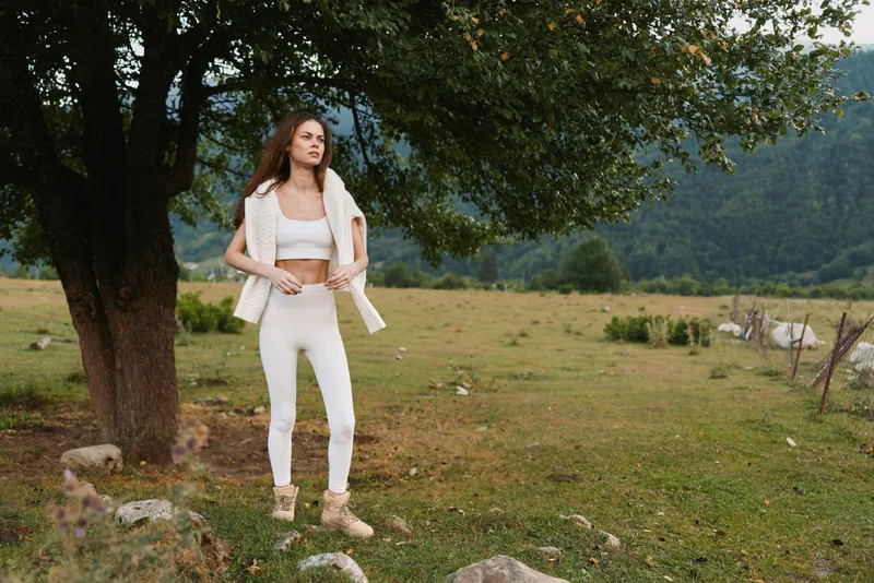 Bright woman outdoors in white attire stands in a sunlit field beside a tree, relaxed pose, natural colors, soft breeze, countryside lifestyle, candid and serene