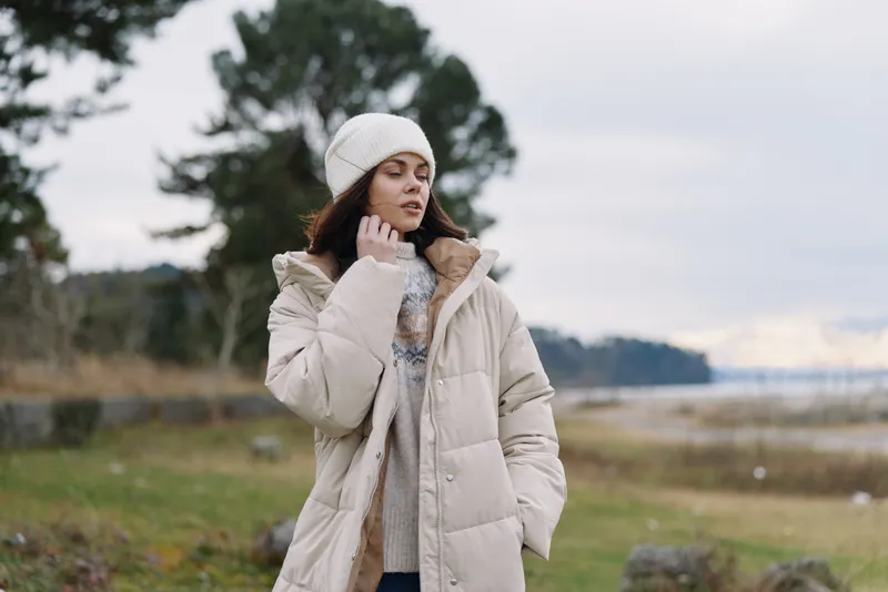 Winter scene with a woman in a beige puffer coat and beanie, standing outdoors near a calm river, with trees in the background, showcasing cozy fashion and a reflective mood.