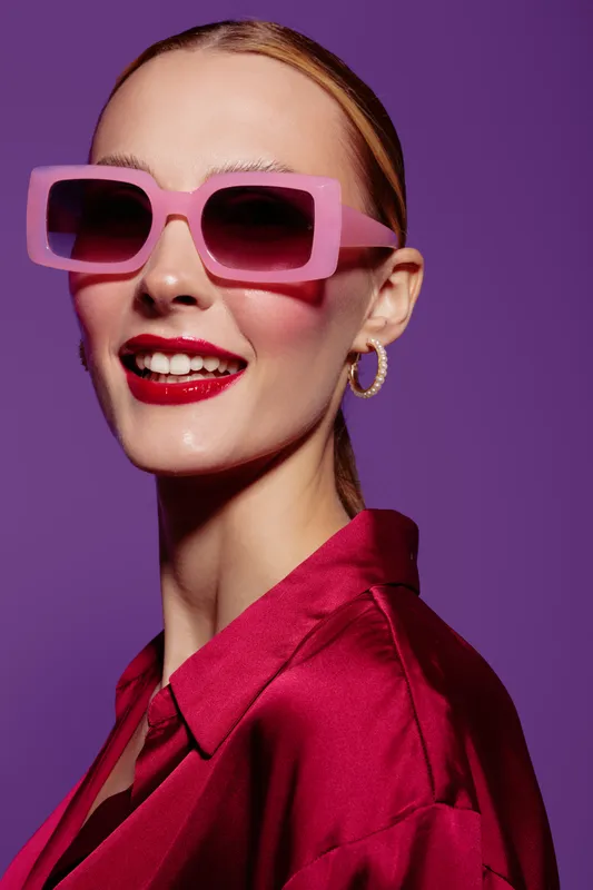 Smiling woman with pink sunglasses and red lipstick wears shiny red blouse. Studio portrait on purple background. Concept of fashion, beauty, style, and confident mood.