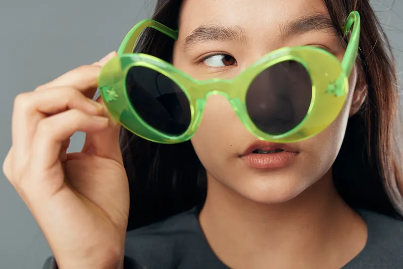 Young woman holding green sunglasses close to her face with focused expression and smooth skin on gray background in studio portrait with casual black shirt and natural makeup.