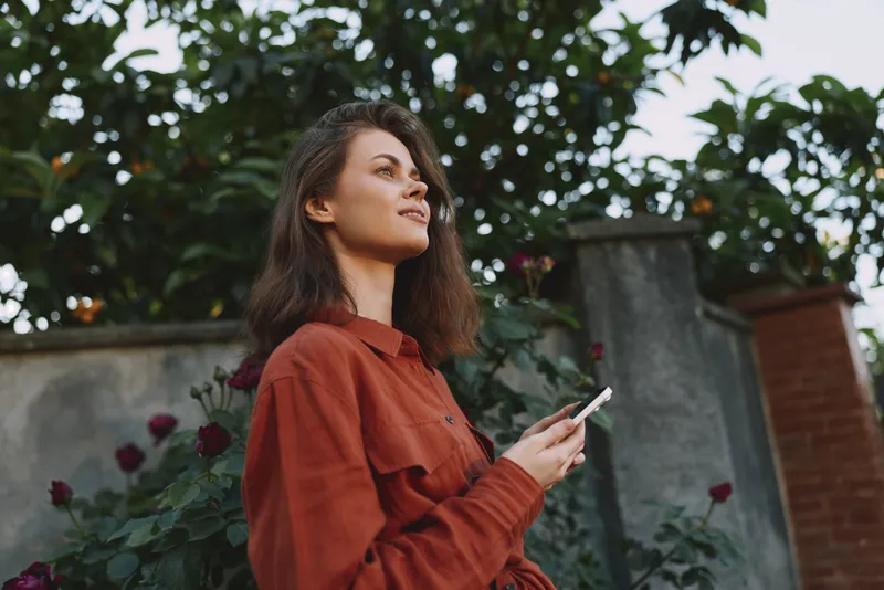Woman in a rust jacket outdoors, holding a smartphone in a garden. Lifestyle portrait emphasizing casual style, relaxation, self expression, and natural light in a sunny daytime scene