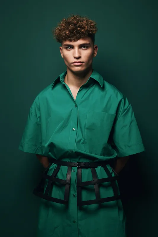 Young man with curly hair wearing oversized green shirt and black harness belt posing against dark green background in studio portrait fashion photography.