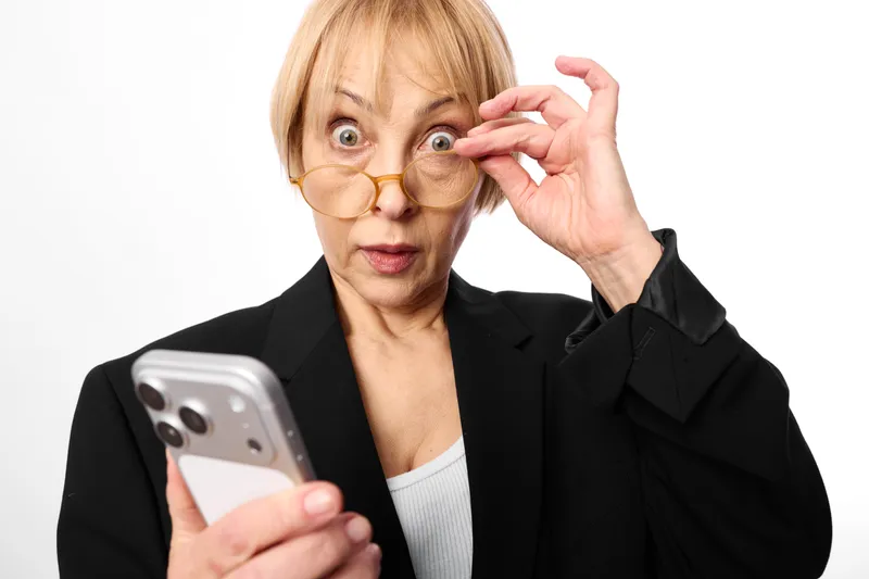Surprised senior woman in studio portrait holding a smartphone, adjusting eyeglasses with one hand, curious expression, black blazer, white background, technology concept