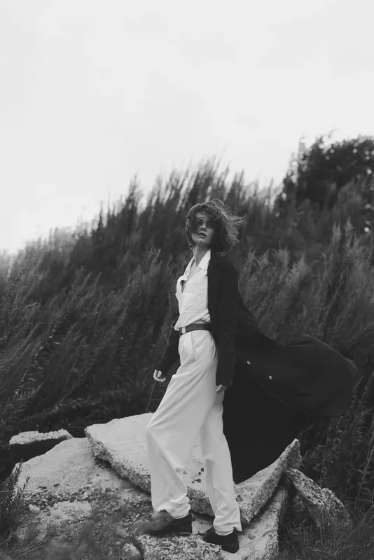 Windy outdoor fashion scene with a woman in a flowing white jumpsuit and long black coat, standing on rocks amid tall grasses, dramatic monochrome style.