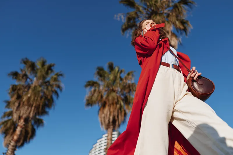 Young woman outdoors wearing a red trench coat over white pants, posing with a bag, sunny sky and palm trees in the background, bold color and confident stance