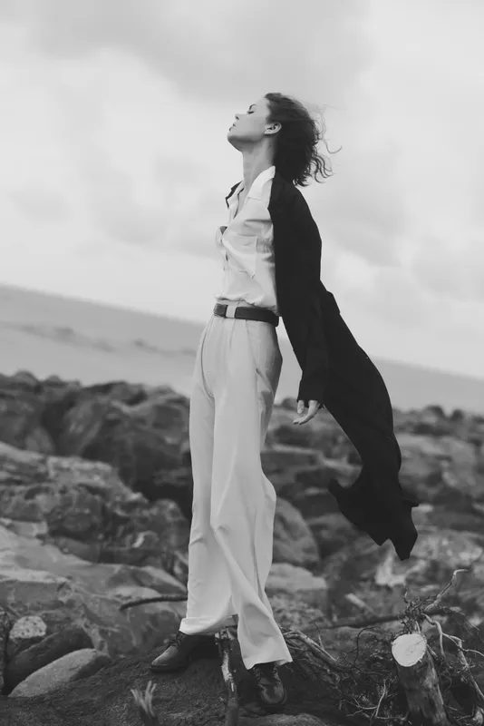 Woman, outdoor, wind, sea, rocks, clothing, standing on a windy shore, flowing pants and blouse billow as waves crash, grayscale fashion mood, dynamic movement, serene coastal atmosphere, editorial