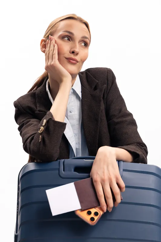 Young woman in business attire leans on blue suitcase holding passport and smartphone. She looks thoughtful and relaxed, concept for travel planning and business trip preparation.
