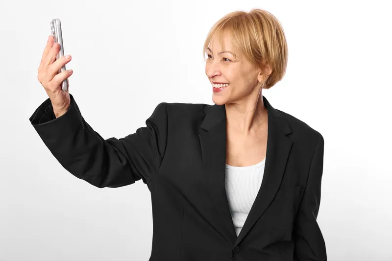 Smiling woman in black blazer on white studio background, holding phone for a selfie, side profile, cheerful expression, clean minimal light, casual confident pose today, well