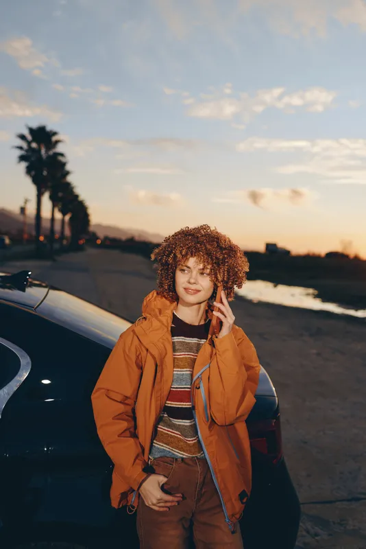 woman lifestyle portrait outdoors at sunset near a car parked on the seaside road curly hair woman wearing an orange jacket and striped sweater relaxed smile looking at camera