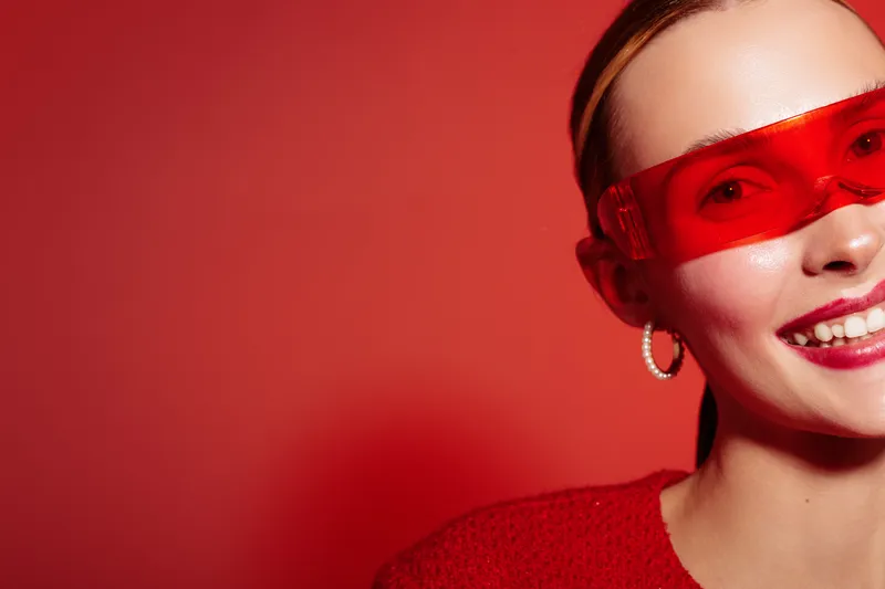 Smiling woman with red transparent glasses and red sweater poses on red background. Close-up portrait with hoop earrings and bright makeup. Concept of fashion, style, and confidence.