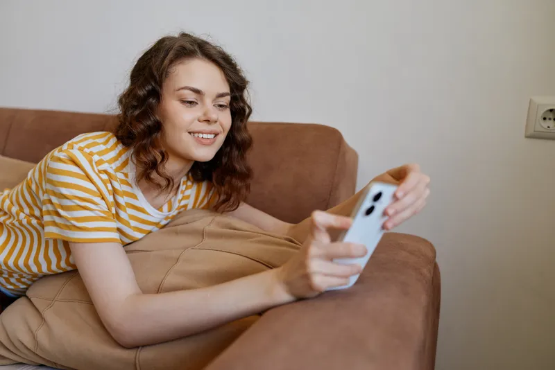 Young woman relaxes on a couch at home, smiling while using a smartphone in a casual striped tee, creating a friendly, intimate moment for a cozy lifestyle scene