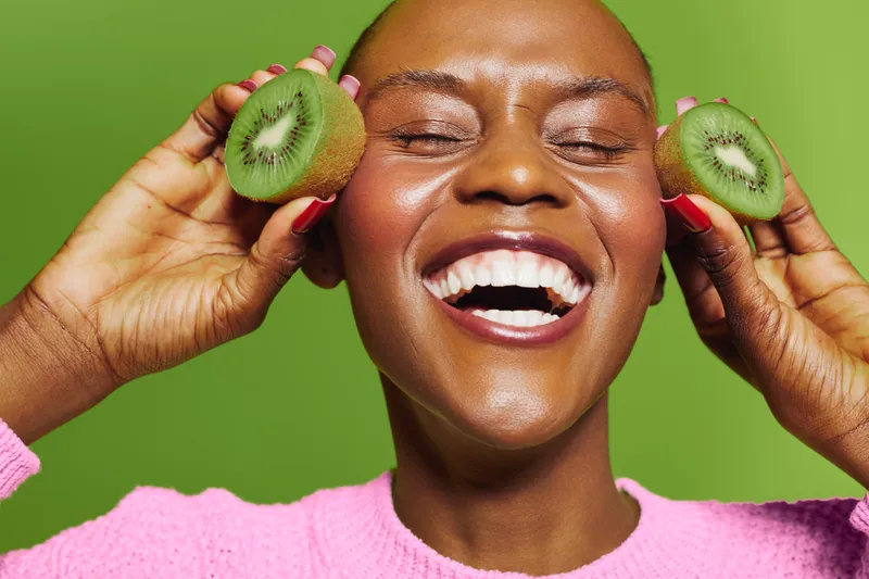 Happy dark skinned African woman with short hair holds two kiwi halves near face laughing joyfully on green background. Concept of healthy lifestyle, fresh fruit, and natural beauty.