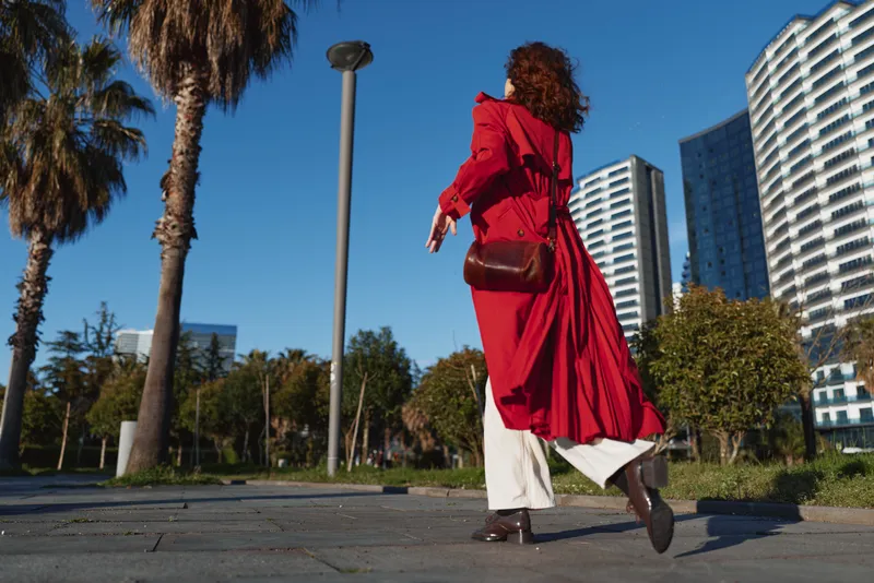 A woman in a red trench coat walks through an urban park with tall buildings behind, creating a dynamic sense of motion, bold fashion, and confident mood on a sunny day.