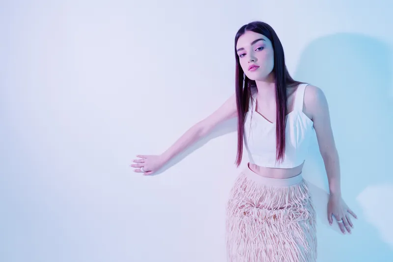 Young woman beauty fashion posing in a white top and textured skirt against a light background with soft pastel lighting. Concept for style, elegance, and modern trends.