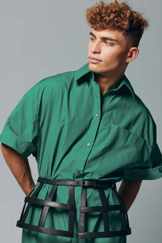 Portrait of young man with curly hair wearing green short sleeve shirt and black belt fashion accessory standing against purple background looking sideways away from camera with serious expression.