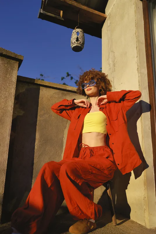 Cheerful woman outdoors in a red jacket and yellow top, leaning against a sunlit wall, urban terrace vibe, relaxed pose, vibrant natural colors, carefree lifestyle moment