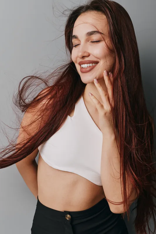 Young woman with long hair and closed eyes smiling in white crop top and black pants. Portrait of happy female with natural makeup and relaxed pose against gray background.