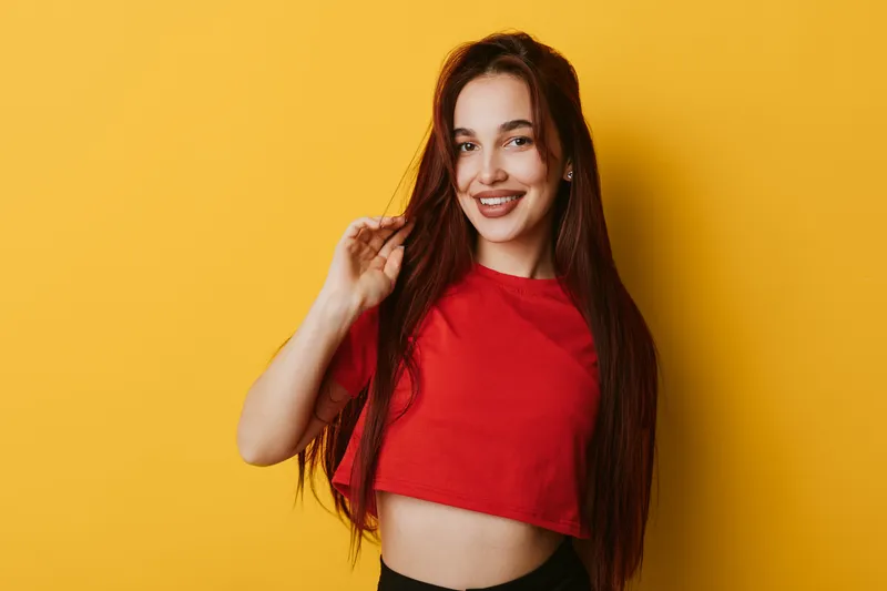 Young woman with long hair smiling and posing in red crop top against yellow background. Casual style, cheerful expression, and confident posture in studio portrait photography.