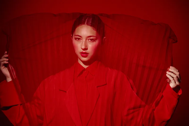 Fashion model dressed in red coat holds large translucent red fan posing with focused face in a creative beauty and fashion concept with dramatic monochrome red lighting.