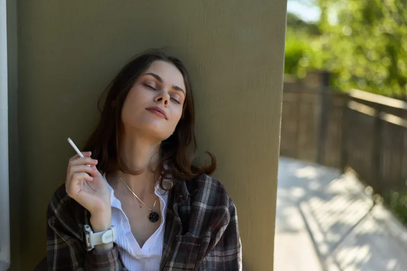 Relaxed woman leans against a sunlit wall, smoking a cigarette with closed eyes and a calm smile, capturing a quiet moment of solo reflection outdoors after a long day