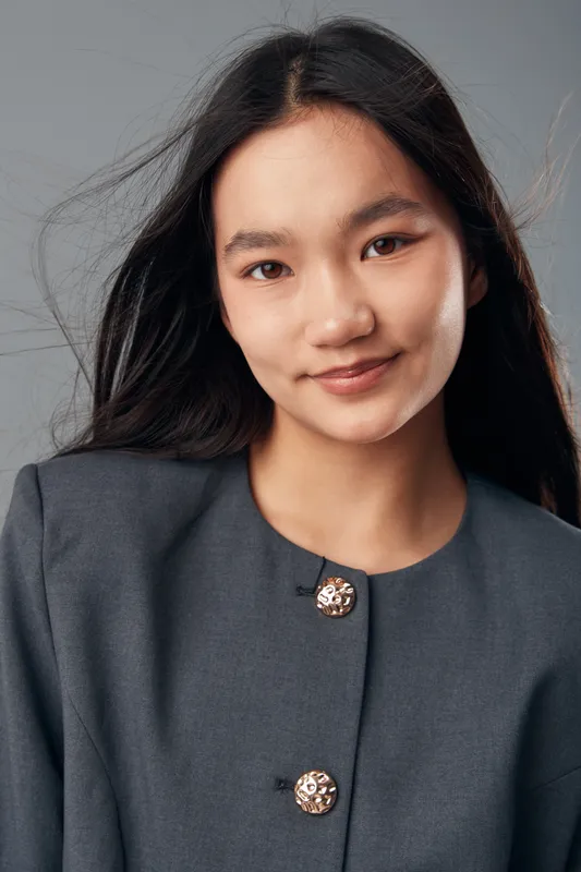 Young woman with long black hair and natural makeup wearing gray jacket posing smiling in studio against neutral background for portrait and fashion concept.