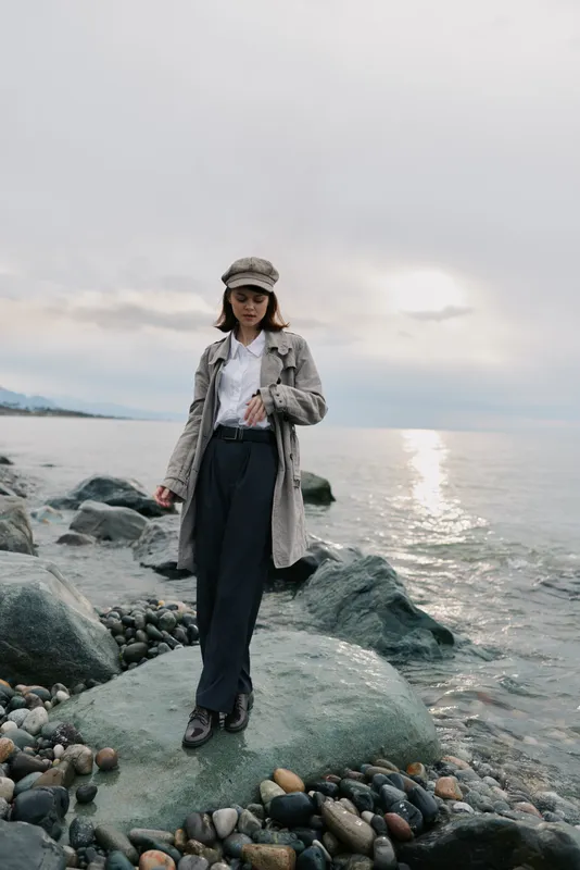 Young woman in casual outfit stands on rocky shore near calm sea under cloudy sky. Concept of nature, fashion, travel, and peaceful outdoor lifestyle by the water.