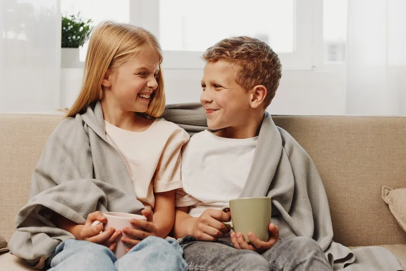 Happy siblings wrapped in cozy blankets sit on a beige sofa sharing warm drinks and smiling at each other in a bright living room. Concept of family bonding, comfort, and childhood moments.