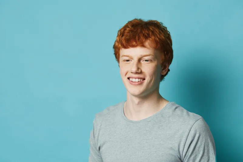 Young man with red hair smiling and wearing gray t shirt on blue background. Portrait of happy male, casual style and positive emotion with copy space.