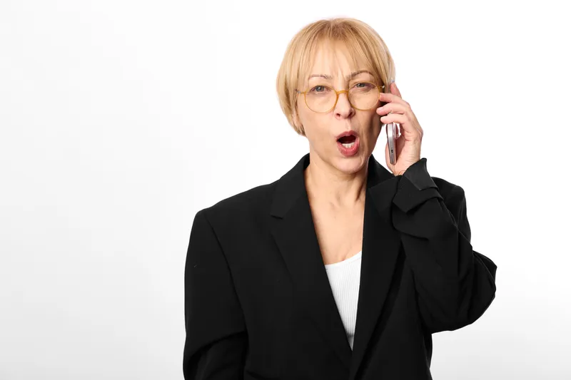 Woman business phone call communication surprised glasses blazer portrait, isolated on white background, adult blonde female speaking on mobile, mouth open, formal office look.