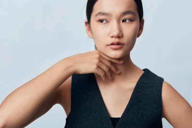 Woman with clear skin and natural makeup posing with hand near neck on light gray background. Beauty model in sleeveless top looking away with thoughtful expression and smooth complexion.