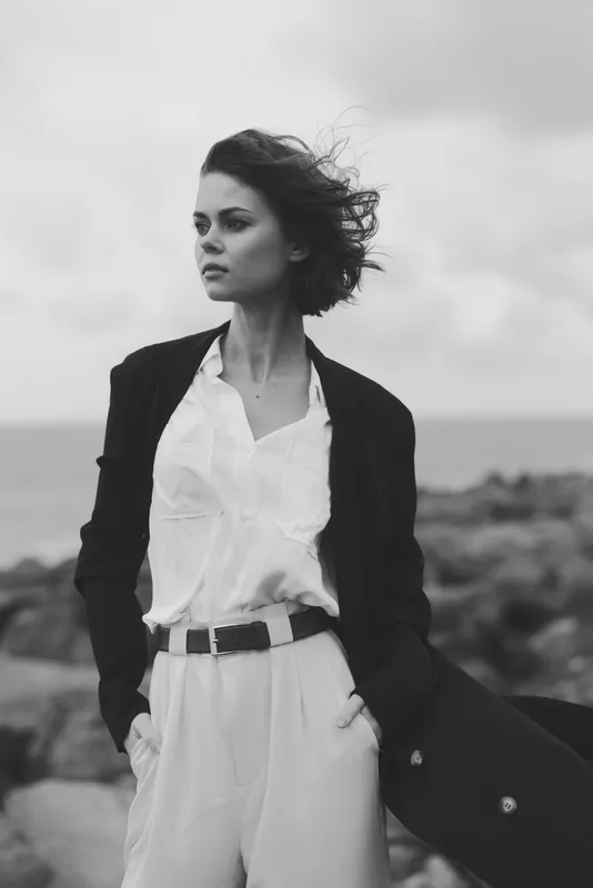 Monochrome image of a confident woman standing outdoors, wind in hair, wearing a white shirt and tailored suit, signaling chic style and resilience by the coast