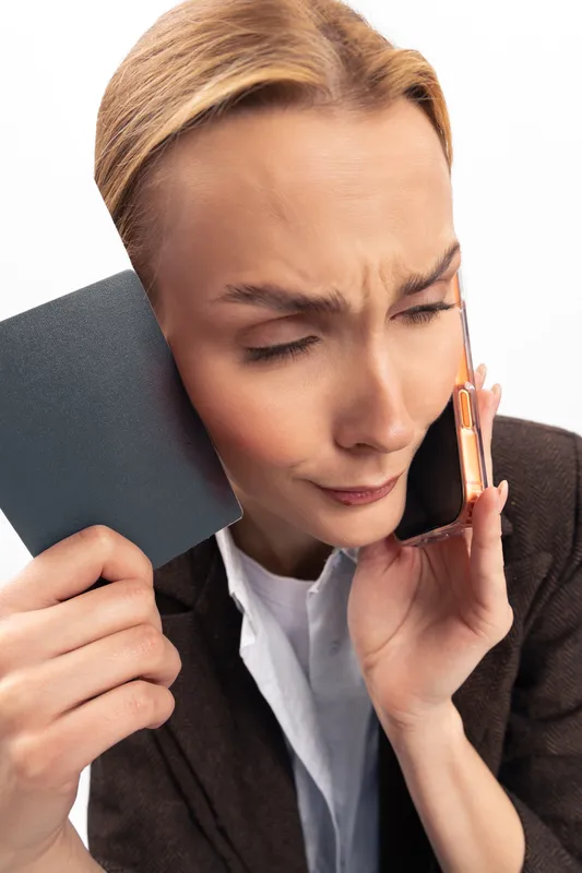 Stressed young woman in dark blazer holds phone to ear and credit card in hand. Facial expression shows frustration during a financial call. Concept of money worries and customer service.