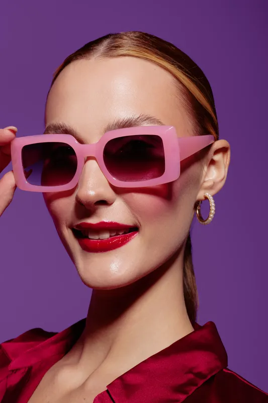 Young woman with pink sunglasses and red lipstick smiles confidently on purple background. Fashion portrait with stylish accessories and vibrant colors for beauty concept.