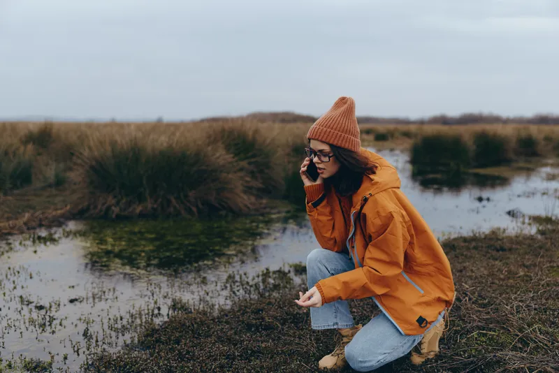 Woman lifestyle outdoors by river in autumn, kneeling on grass, wearing orange hat, jacket and sunglasses, calm water nearby, relaxed travel mood in nature landscape