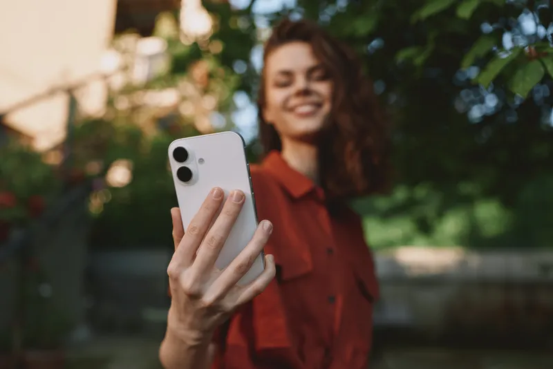 Woman lifestyle shot capturing a smiling woman outdoors taking a selfie with a smartphone, casual red shirt, lush greenery, candid portrait in a sunny park setting for stock