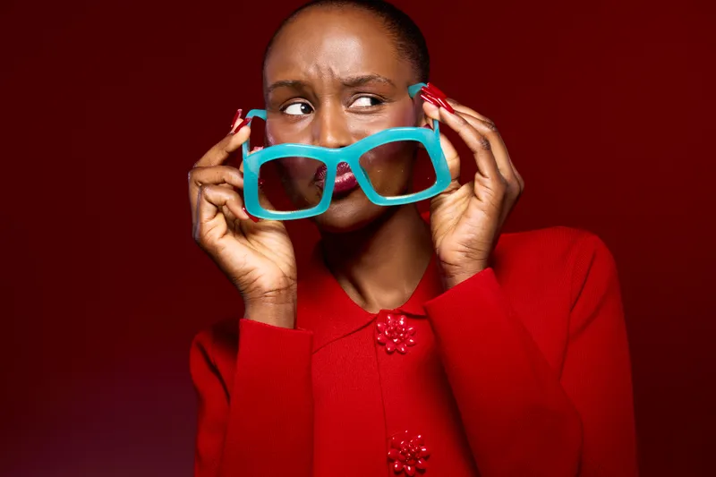 Portrait of a thoughtful black woman with dark skin holding large turquoise glasses near her face. She wears a red sweater against a red background. Concept of fashion and style.