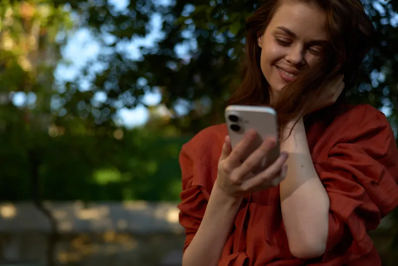 Smiling woman outdoors looking at smartphone, casual red top, natural greenery backdrop. Concept of lifestyle, wellness, and social connection. Warm, relaxed mood