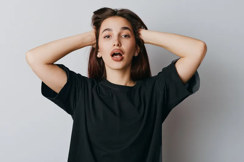 Young woman with long hair in black t shirt posing with hands behind head and mouth slightly open on white background for emotion, expression and casual style portrait.