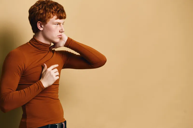 Redhead man with thoughtful expression wearing brown turtleneck standing against beige wall looking sideways with one hand on chest and the other near neck in casual pose.