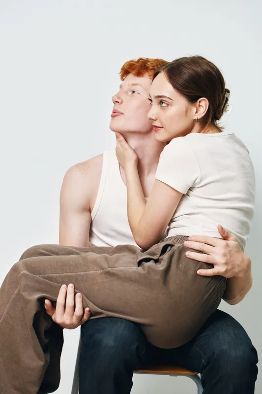 Young man and woman posing in studio, woman sitting on man’s lap with gentle touch and close connection, casual clothes and neutral background for relationship or friendship.