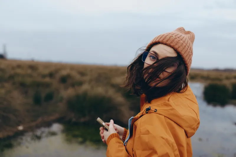 Woman outdoors in an orange coat and knit hat by a river landscape, scarf covering face, holding smartphone, winter lifestyle travel mood, soft overcast light, shallow depth of field, nature