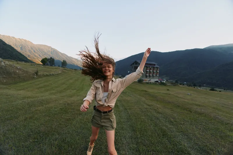 A young woman in casual clothes joyfully runs across a green field with mountains and a building in the background during daylight. Lifestyle and outdoor activity concept.