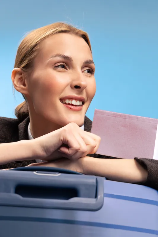 Smiling woman holding passport and leaning on a blue suitcase with a bright blue sky background. Concept of travel, vacation, and holiday planning with positive emotions.