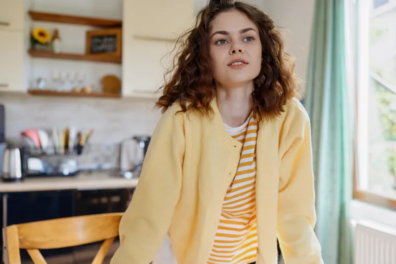 Young woman in a bright yellow cardigan stands in a cozy home kitchen, natural light on her face, relaxed lifestyle moment, casual portrait of everyday life in a warm interior