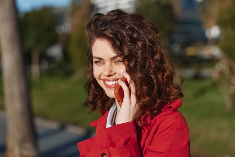 Smiling young woman with curly hair talks on a smartphone outdoors, wearing a red jacket in a sunlit park setting, expressing confidence and casual conversation.